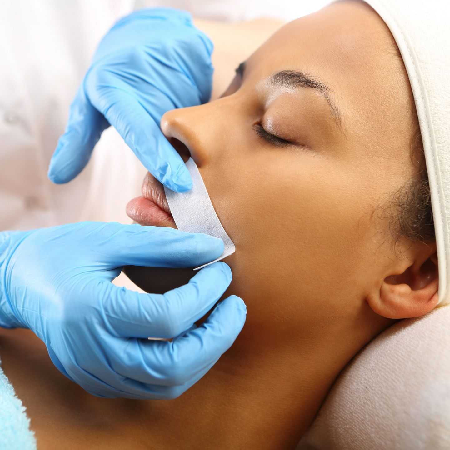 Person having upper lip hair removed with a waxing strip applied by a professional wearing gloves.